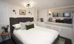Modern bedroom with a grey upholstered bed, white bedding, a wall-mounted horse painting, built-in shelving, and a window seat adorned with cushions and decorative plants.